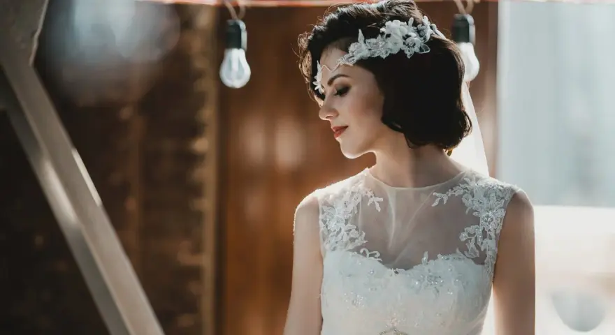 Panna młoda z krótkimi włosami w stylu boho, ozdobionymi kwiatową opaską, w białej sukni z koronkowymi detalami.
