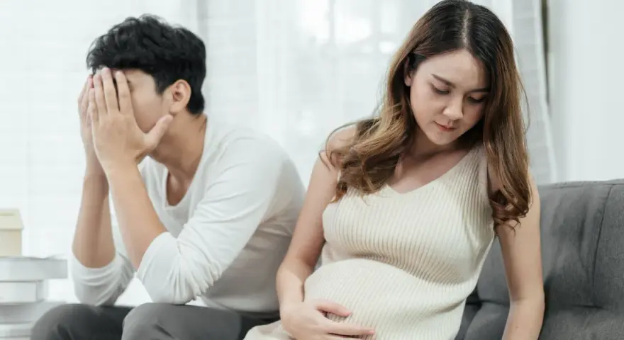 Ein Mann mit verdecktem Gesicht sitzt abgewandt von einer schwangeren Frau. Die Szene deutet auf eine Trennung in der Schwangerschaft hin, die mit Kummer verbunden ist.