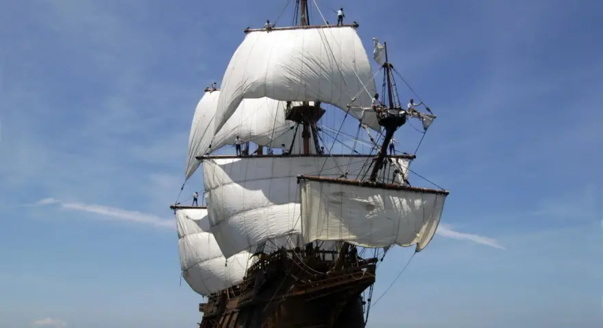 Un majestueux galion espagnol navigue sous un ciel bleu, ses voiles blanches gonflées par le vent.
