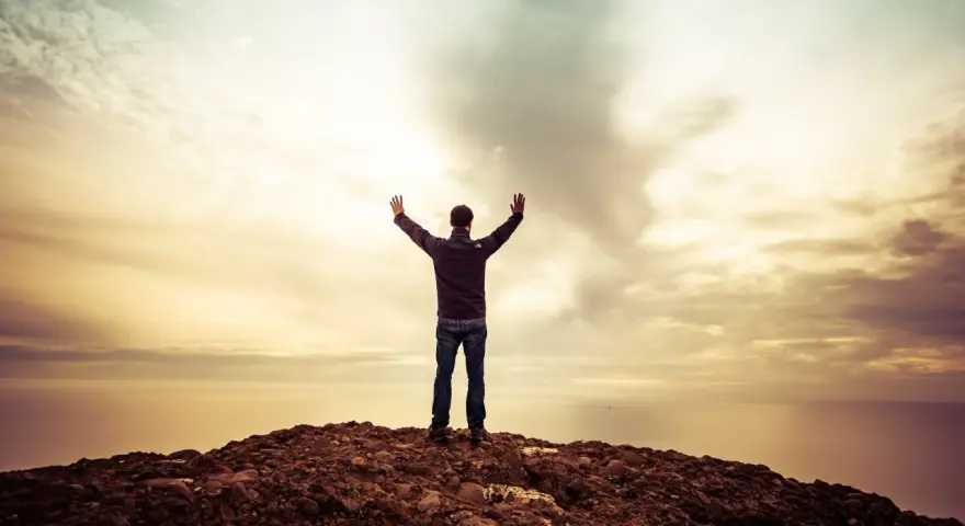 Mann mit erhobenen Armen auf einem Felsen, blickt auf das Meer. Ein Gefühl des aaronitischen Segens liegt in der Luft.