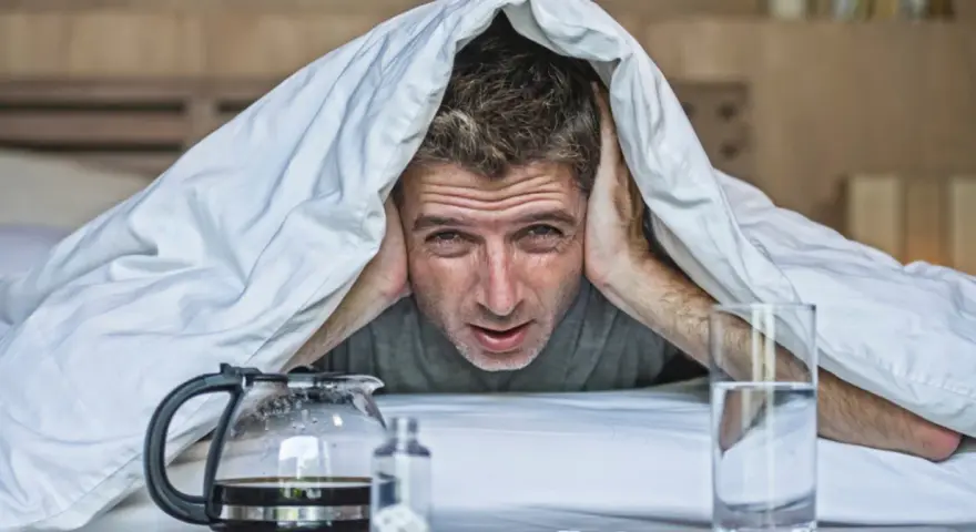 Mężczyzna pod kołdrą z rękami na uszach, z kawą i wodą obok. Wygląda na to, że ma uczucie kaca bez alkoholu.