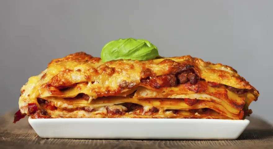 Una porción generosa de lasaña de carne, con capas de pasta, salsa y queso dorado, coronada con una hoja de albahaca fresca.