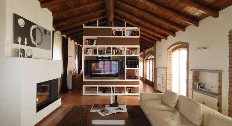 Accogliente soggiorno mansardato con soffitto in legno a vista, camino moderno e libreria centrale. Idee mansarde in legno per un ambiente caldo.