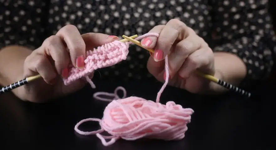 Händerna stickar en rosa v-ringning med ljusa trästickor. En garnnystan ligger bredvid.