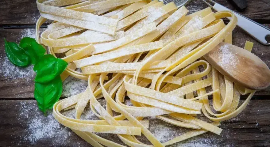 Świeży, domowy makaron bez glutenu, posypany mąką, z listkami bazylii i drewnianą łyżką.