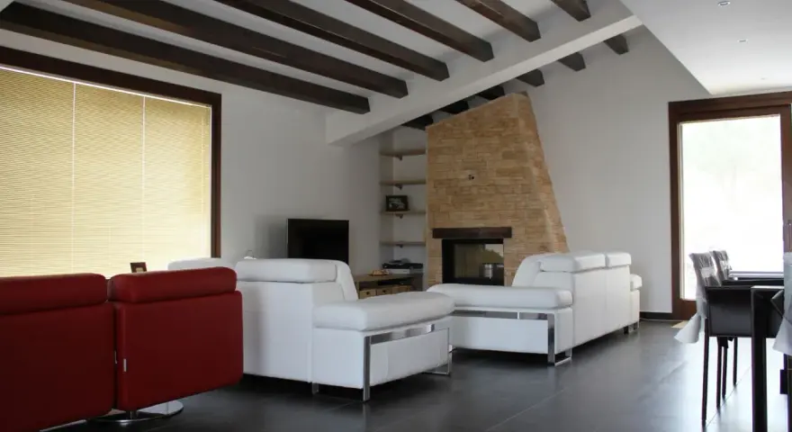 Soffitto con travi in legno scuro, un camino in pietra e divani bianchi. Ecco come fissare travi in legno al soffitto per un effetto rustico.