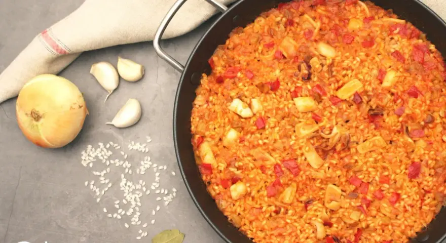 Arroz con calamares en paellera, con ingredientes como cebolla, ajo y pimientos. Un plato delicioso y lleno de sabor.