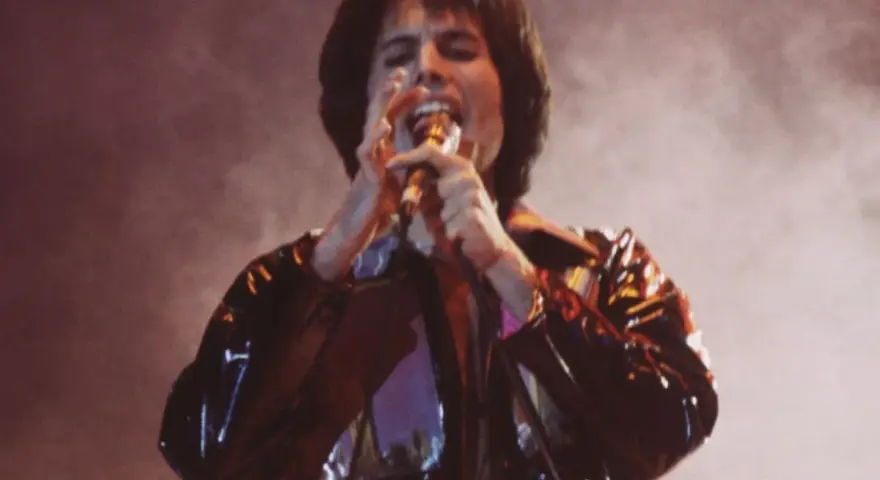 Freddie Mercury śpiewa z pasją, czując, że chce się uwolnić. Błyszcząca kurtka odbija światła sceny.