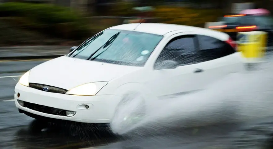 Auto bianco in movimento su strada bagnata, con un'enorme spruzzata d'acqua che indica l'aquaplaning cos'è.