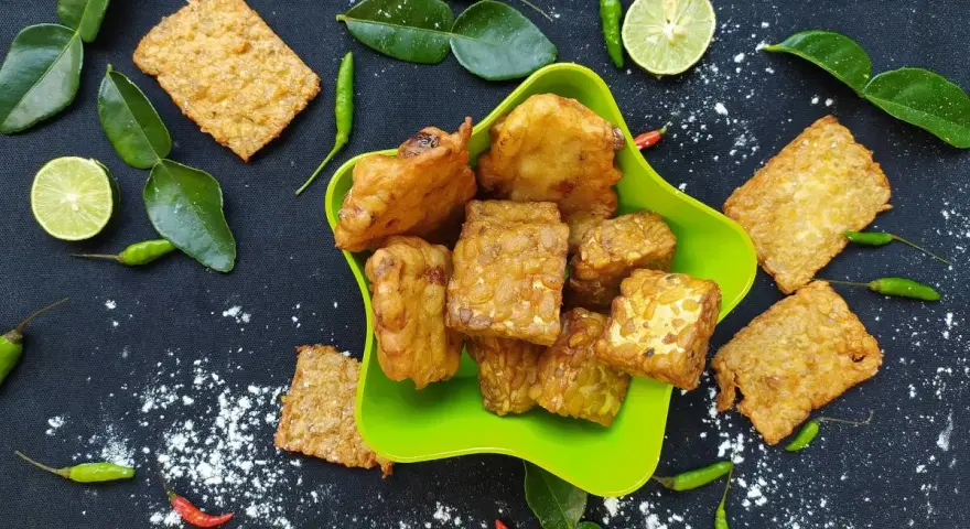 Knusprig gebratener Tempeh, ein fermentiertes Sojaprodukt, liegt in einer grünen Schale. Dazu garnieren Limettenscheiben, grüne Chilis und Blätter.