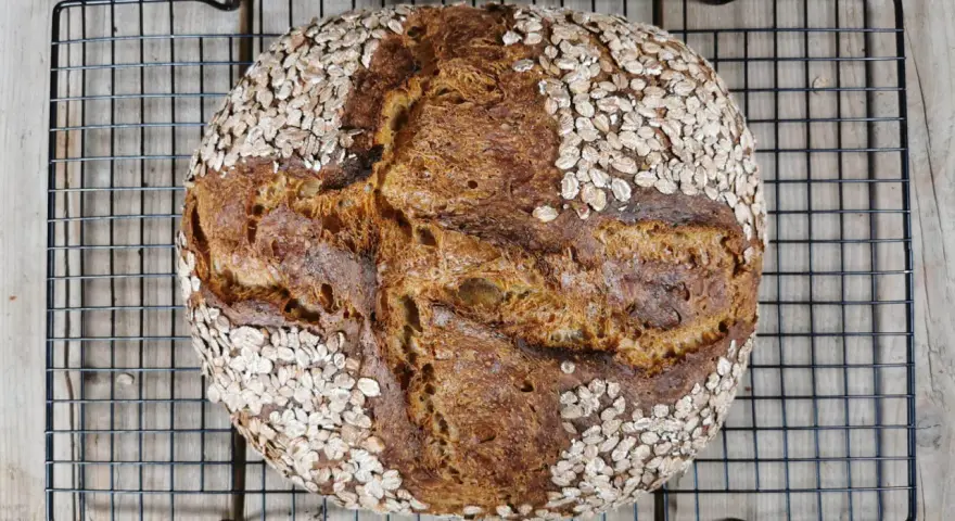 Ein rustikales Porridge Brot mit Haferflockenkruste, das auf einem schwarzen Gitter auf Holzdielen abkühlt.