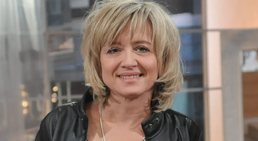 Uśmiechnięta Małgorzata Bogdańska w czarnej ramonesce, z blond włosami i delikatnym makijażem.