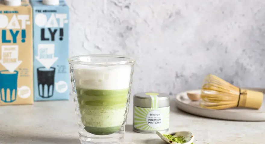 Warstwowy napój matcha z mlekiem kokosowym w szklance z podwójnymi ściankami. Obok puszka z matchą i bambusowa miotełka.