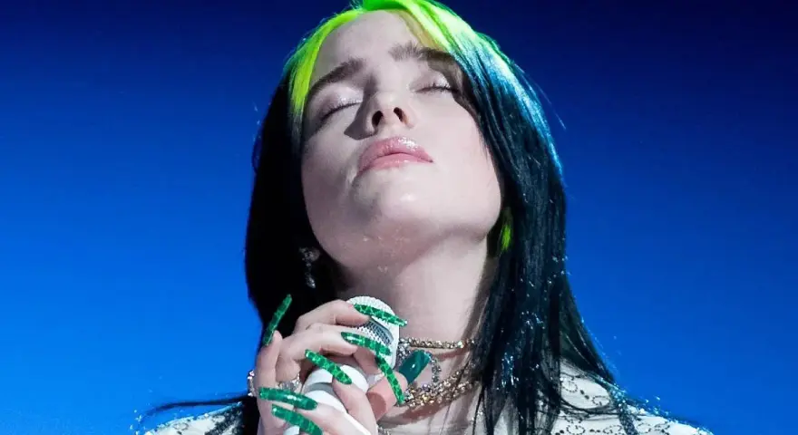Piosenkarka Billie Eilish z zamkniętymi oczami śpiewa do mikrofonu, jej zielone pasemka włosów lśnią.