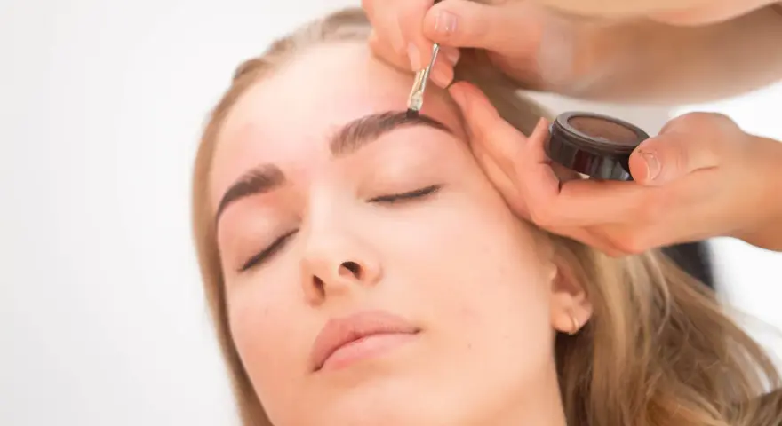Profesjonalne malowanie brwi krok po kroku. Makijażystka precyzyjnie modeluje łuk brwiowy za pomocą pędzelka i cienia.