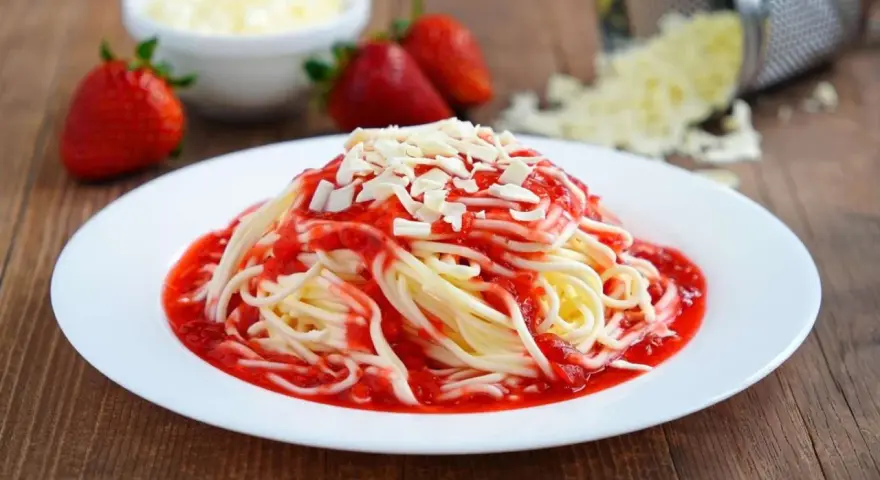 Ein Teller mit Spaghetti Eis, garniert mit Erdbeersauce und weißen Schokostückchen. Frische Erdbeeren und geriebene Schokolade im Hintergrund.