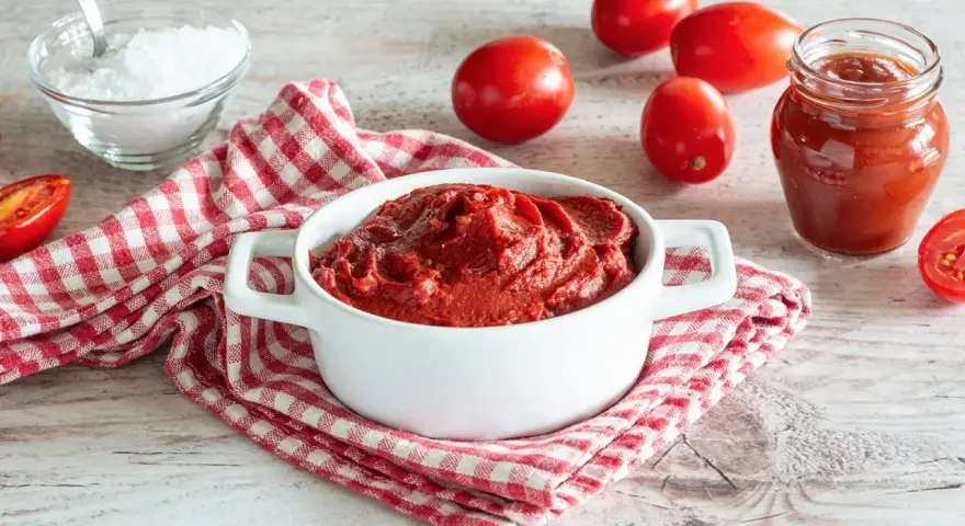 Un bianco contenitore con un denso macerato di pomodoro, circondato da pomodori freschi, sale e un barattolo di salsa.