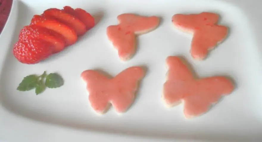 Zarte Erdbeer-Kekse in Schmetterlingsform, dekoriert mit rosa Zuckerguss, neben frischen Erdbeerscheiben und einem Minzblatt.