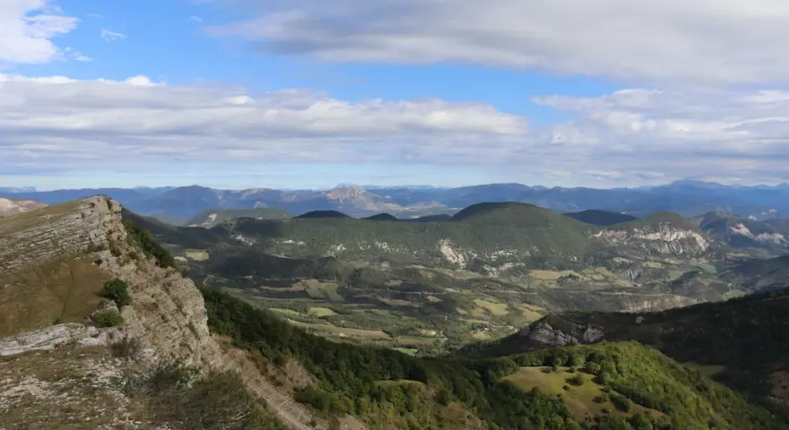 Vue panoramique depuis le col de Perty, avec des montagnes verdoyantes et un ciel bleu parsemé de nuages.