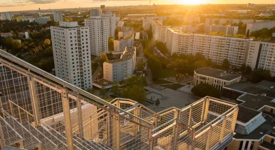 Blick von einer Treppe auf den Skywalk Marzahn bei Sonnenuntergang. Hohe Wohnhäuser und grüne Flächen prägen die Szenerie.