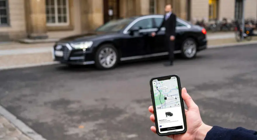 Hand hält Smartphone mit Uber-App, die eine Fahrt mit uber premium cars anzeigt. Ein Fahrer steht neben einem schwarzen Audi.