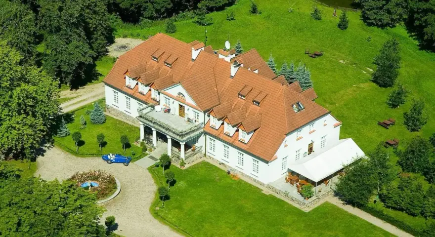 Dwór w Bychowie otoczony zielenią, z niebieskim kabrioletem przy różanym klombie.