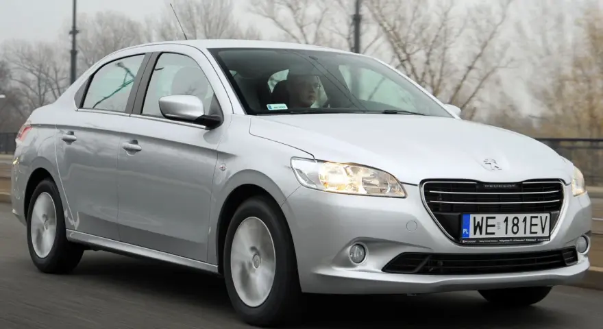 Srebrny Peugeot 301 jedzie po drodze, za kierownicą siedzi mężczyzna w okularach.