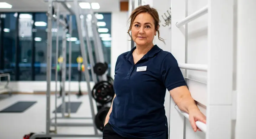 En kvinna i träningskläder står i ett gym. Hon har en namnskylt på sin tröja. I bakgrunden syns träningsredskap, som kan användas för att stärka underbenets muskler.