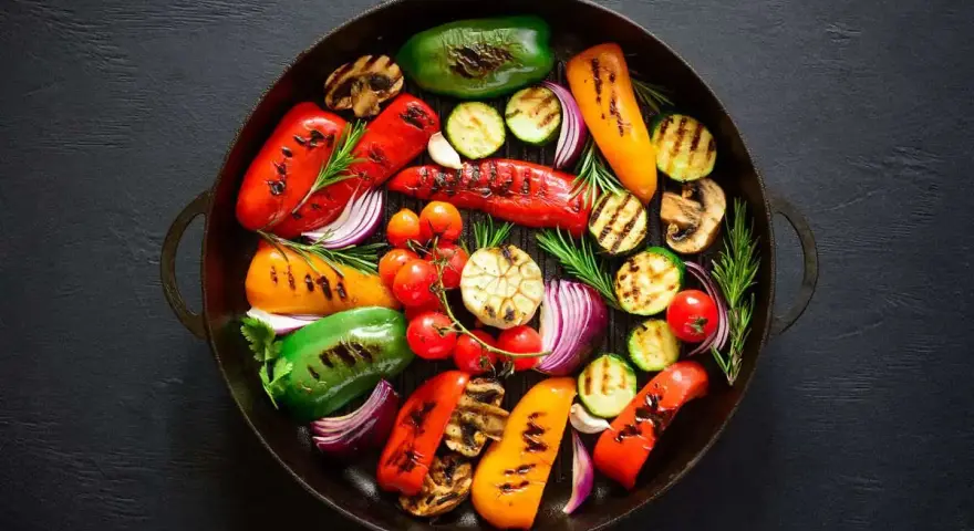 Un assortimento colorato di verdure grigliate, tra cui peperoni, zucchine, pomodorini, cipolle rosse e funghi, disposte in una padella scura.