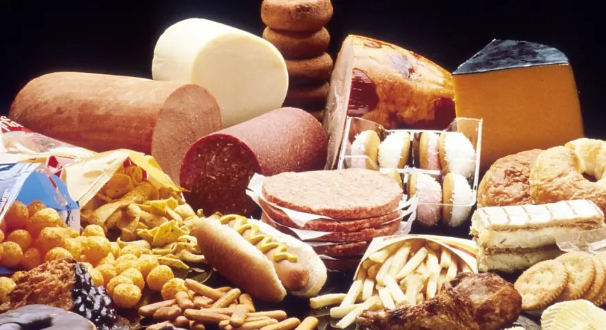 Stół ugina się pod ciężarem przetworzonej żywności: wędliny, sery, frytki, słodycze, ciastka, pączki, hamburgery i chipsy – to wszystko zawiera tłuszcze trans.