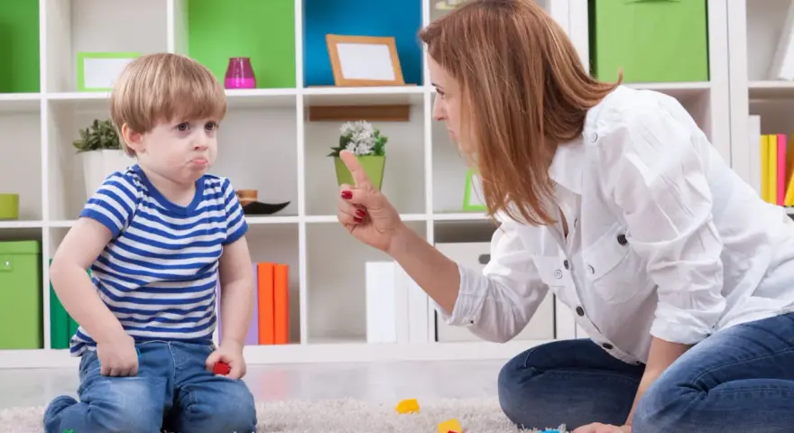 Mama karci synka za kłamstwo. Chłopiec ma smutną minę, a na dywanie leżą klocki.