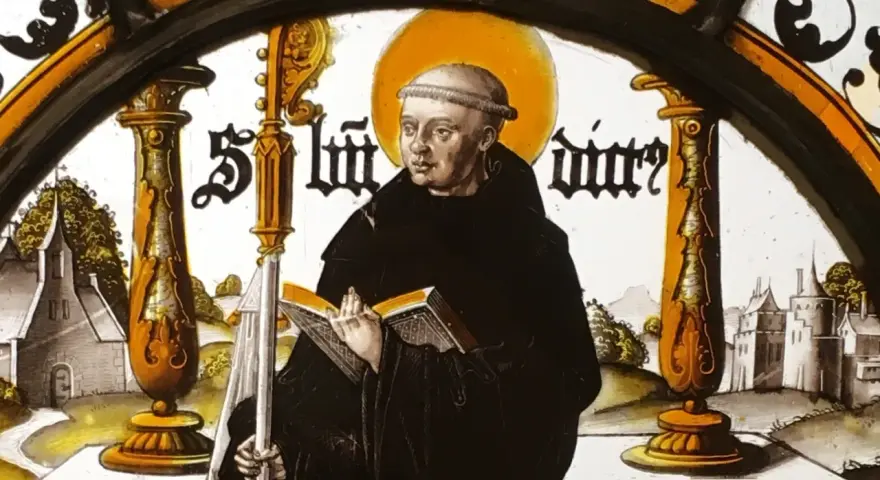 Stained glass depicting St. Benedict the Abbot, holding a book and staff, with a monastery in the background.