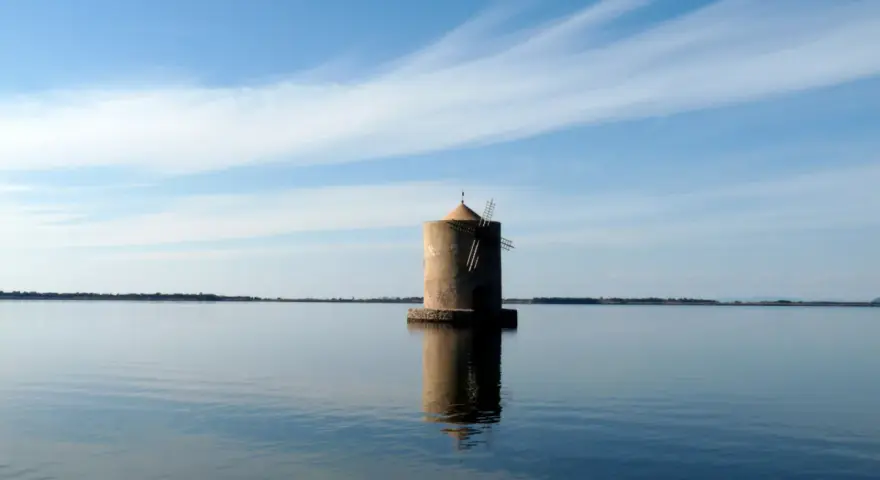 Mulino solitario nella laguna di Orbetello, riflesso sull'acqua calma sotto un cielo azzurro con nuvole sottili.