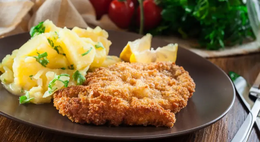 Kotlet schabowy w panierce z ziemniakami i cytryną na brązowym talerzu.