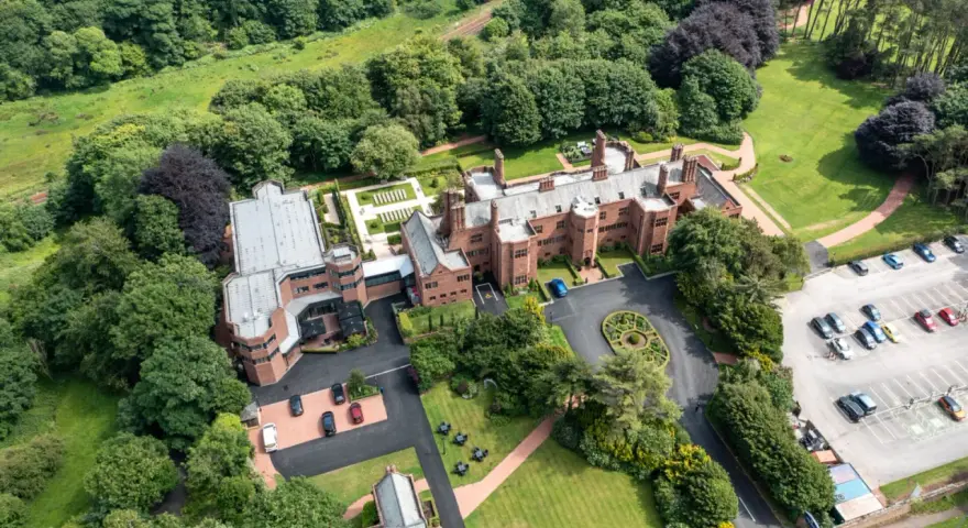 Aerial view of the abbey guest house, a grand brick building surrounded by lush green trees and manicured lawns. Cars are parked in a lot.