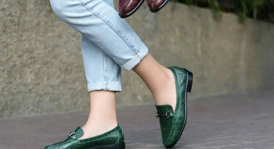 Un mocassino verde con abito elegante, perfetto per un look sofisticato. Un altro paio marrone è tenuto in mano.