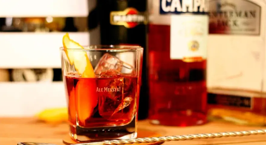 Klasyczny drink z pomarańczą i lodem. Wybór między Negroni czy Boulevardier? Na stole widać butelki Martini, Campari i Jack Daniel's.