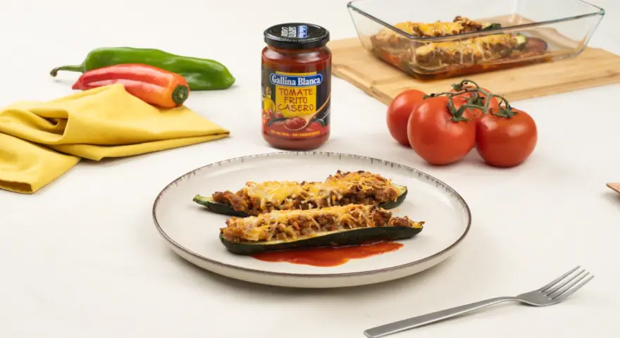 Dos calabacines rellenos de carne y gratinados con queso, servidos en un plato con tomate frito.
