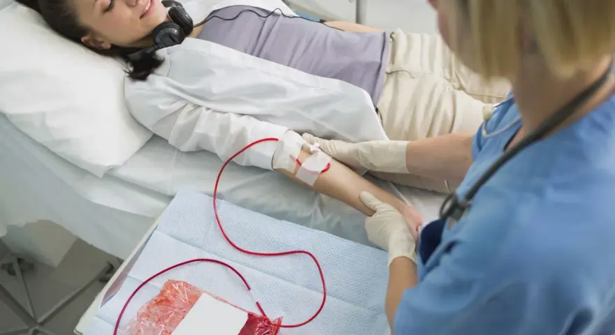 Kobieta leży w łóżku szpitalnym, słuchając muzyki przez słuchawki. Pielęgniarka wykonuje przetaczanie krwi, podłączając worek z krwią do jej ręki.
