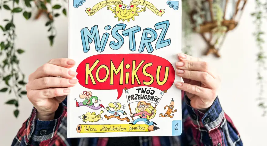 Okładka książki "Mistrz Komiksu" z rysunkami postaci i słońca, trzymana przez ręce.