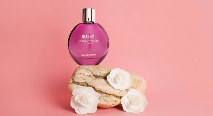 Różowa butelka perfum JFenzi Mon Amie Metamorphoze Women na kamieniu z białymi kwiatami.