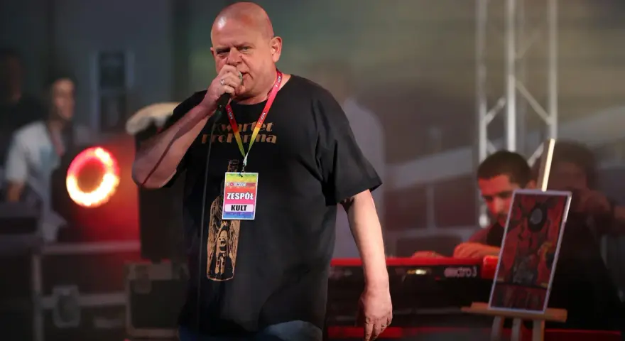 Kazik Staszewski śpiewa na koncercie, ubrany w czarną koszulkę z napisem "Kwartet Pro Forma" i identyfikatorem "Zespół KULT".