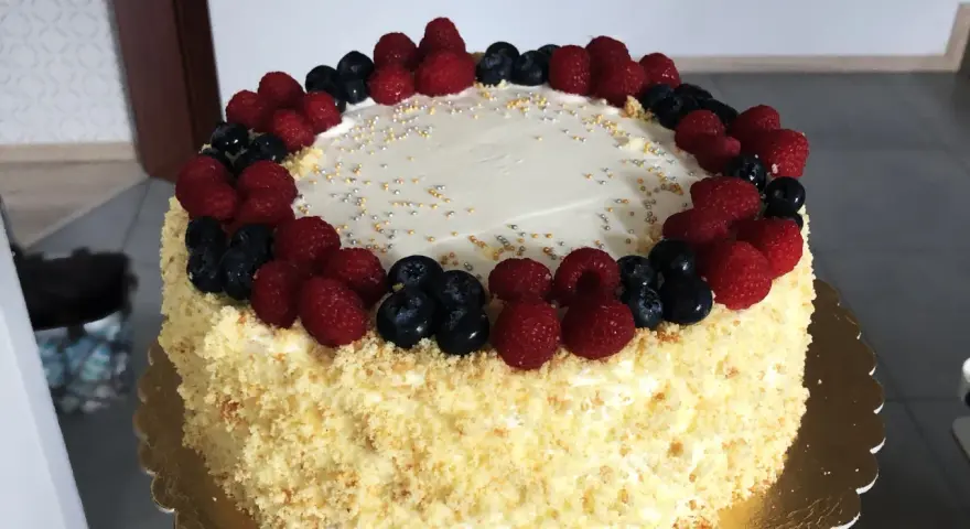 Pyszny tort śmietankowo-malinowy z mascarpone, ozdobiony świeżymi malinami i borówkami, posypany złotymi i srebrnymi kuleczkami.