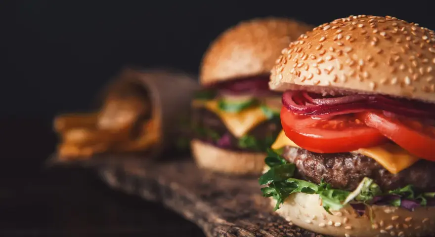 Dwa soczyste burgery z serem, pomidorem i czerwoną cebulą na drewnianej desce, obok frytki.