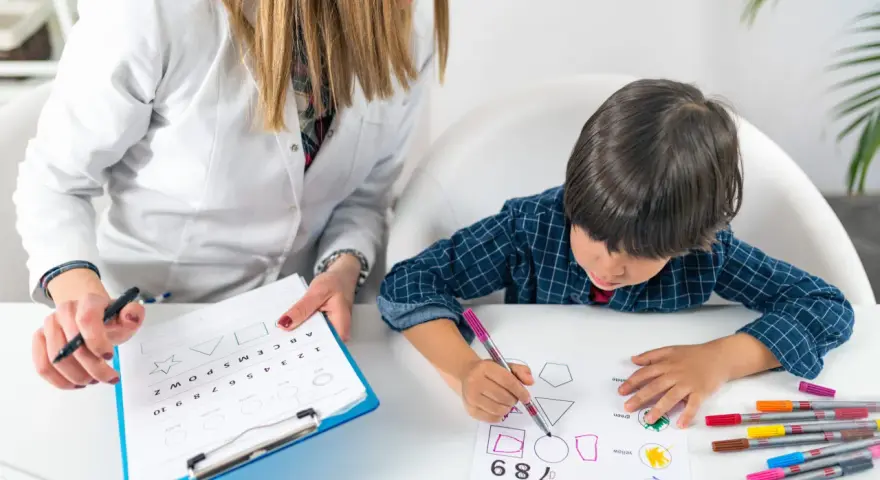 Bambino impegnato in un test per l'autismo, colora forme geometriche guidato da un'insegnante.