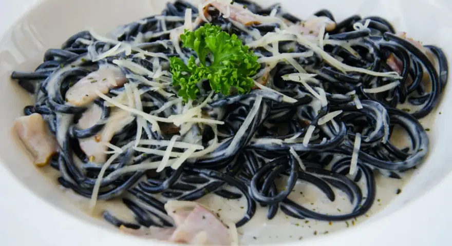 Czarny makaron spaghetti w kremowym sosie, posypany startym parmezanem i udekorowany gałązką natki pietruszki.