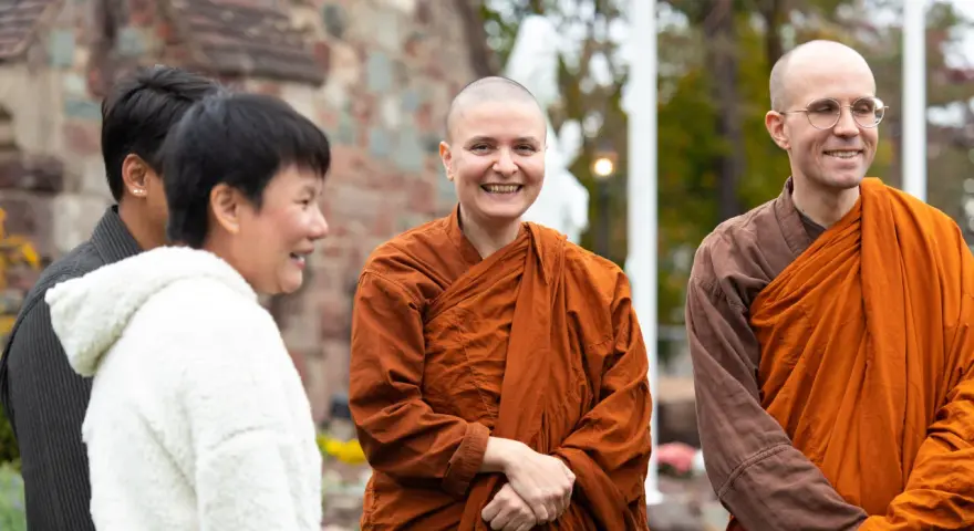 A smiling Buddhist monk in orange robes stands with others outside a church.