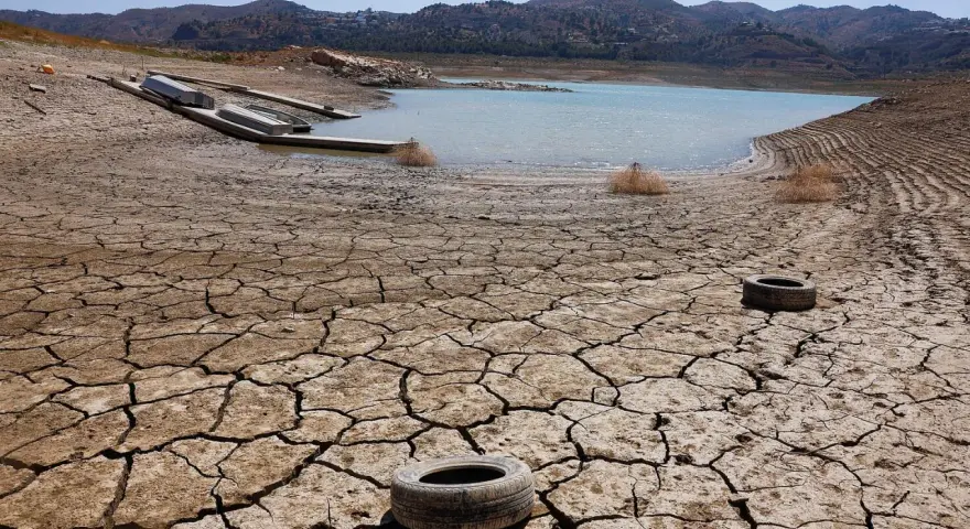 Seca tierra agrietada con un lago bajo y barcos varados. Neumáticos abandonados sugieren una contaminación natural y humana.