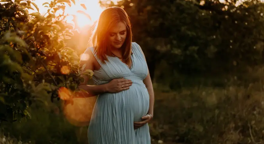 Piękne zdjęcia w ciąży. Kobieta w błękitnej sukience trzyma dłonie na brzuchu, otoczona ciepłym światłem zachodzącego słońca.