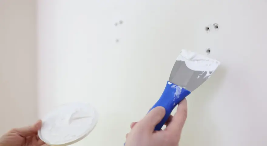 Persona aplica masilla en una pared blanca con una espátula azul para reparar agujeros.
